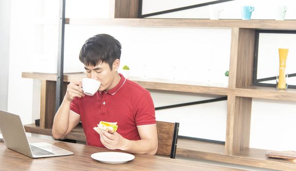 Азиатский улыбающийся молодой человек в обычной красной футболке наслаждаться завтраком, есть сэндвич и работать с ноутбуком, молодой человек приготовления пищи и напитков в лофт стиле кухни
 - Фото, изображение