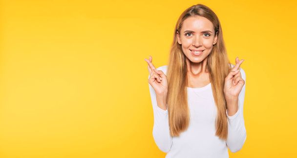 Make a wish and believe in miracle. Happy young cute woman with crossed fingers isolated on yellow background - Fotó, kép