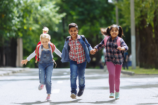 Возвращайся в школу. Смешанная расовая группа счастливых учеников начальной школы с рюкзаками, бегущими за руки на улице. Концепция начального образования
. - Фото, изображение