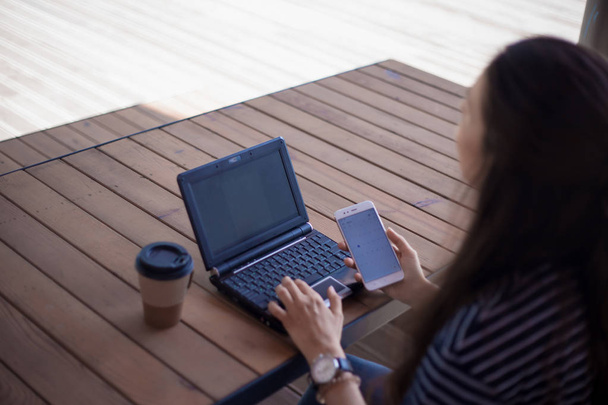 Brunette meisje met een groot polshorloge, half-gedraaid, werkt op een laptop staande op tafel. In de buurt is een smartphone. - Foto, afbeelding