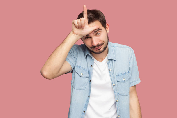 bearded young man in blue casual style shirt showing loser sign on forehead and looking at camera with sad face on pink background - 写真・画像
