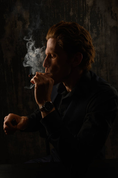 Confident casual young man and smoking cigarette. Studio portrait against dark background. - Фото, изображение
