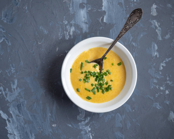 Σούπα από πουρέ πατάτας με λαχανικά και τυρί με πράσινα κρεμμύδια σε μια βαθιά άσπρη πλάκα, μια κουταλιά της σούπας σε γκρίζο φόντο, κορυφή θέα - Φωτογραφία, εικόνα
