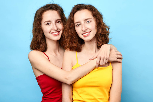 Pleased awesome twins wearing t-shirts hugging each other and looking at the camera over blue background - Фото, изображение