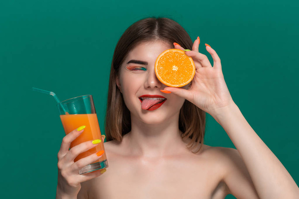 オレンジジュースを持ち、オレンジの半分を噛む明るい色のメイクアップを持つ美しい若い女性のクローズアップ肖像画 - 写真・画像