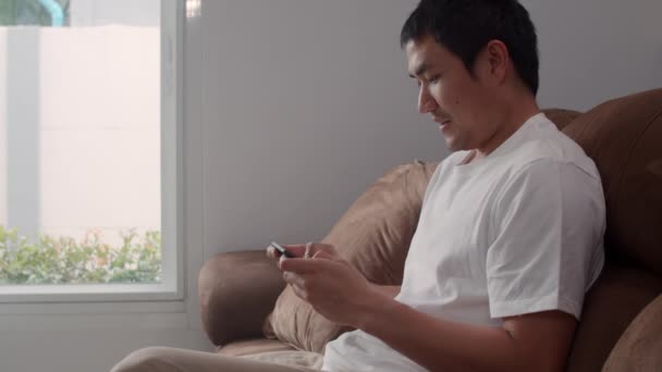 Genç Asyalı adam cep telefonu kullanıyor oturma odasında televizyon oyunu oynuyor, erkek kendini mutlu hissediyor evdeki kanepede uzanmış dinlenme zamanını kullanıyor. Erkekler ev kavramında oyun oynar rahatlar.. - Video, Çekim