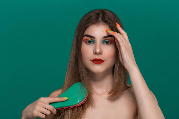 Porträt einer schönen jungen Frau mit leuchtend farbigem Make-up, das die Haare mit einem grünen Pinsel kämmt - Foto, Bild