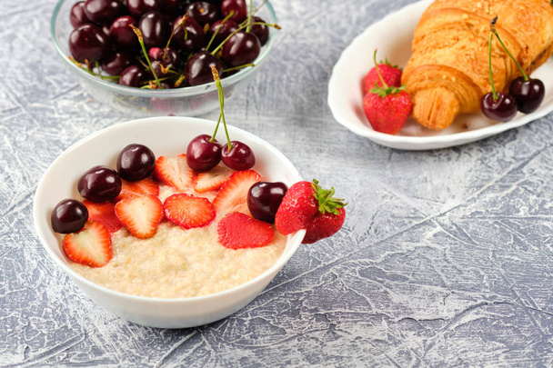 weiße Schüssel Brei mit Erdbeeren und Kirschen, Croissants und Knäckebrot mit Käse und Erdbeeren auf grauem Hintergrund - Foto, Bild