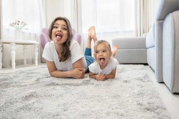 beautiful young brunette woman with cute baby girl wearing jeans and white t-shirts lying on the floor in bright livingroom showing togues - Φωτογραφία, εικόνα