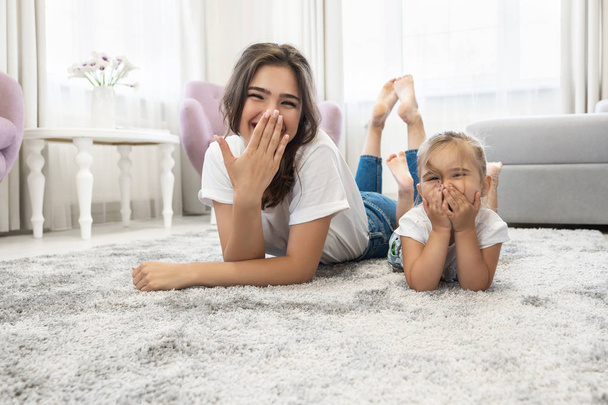beautiful young brunette woman with cute baby girl wearing jeans and white t-shirts lying on the floor in bright livingroom laugh closing their mouths - Photo, image