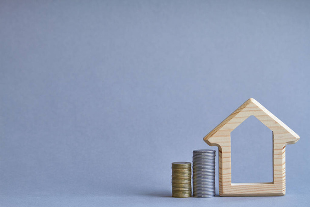 Uma estatueta de madeira da casa com duas colunas de moedas próximas em fundo cinza, o conceito de comprar ou alugar um edifício, foco seletivo
 - Foto, Imagem