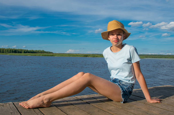the girl in the hat sits on the pier, against the blue sky and the lake, gets a sun bath - Foto, immagini