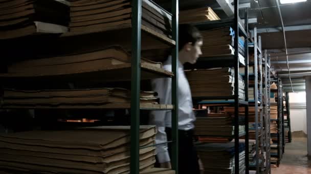 Een tiener in een wit overhemd is op zoek naar een kleine jongen tussen de planken met oude boeken. Donkere kamer in de bibliotheek of archiefruimte met planken van oude literatuur en documenten. - Video