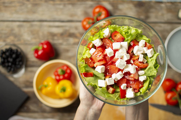 hands holding a vegetarian salad in a glass plate, sliced healthy food ingredients on a wooden table background, close-up - Photo, Image