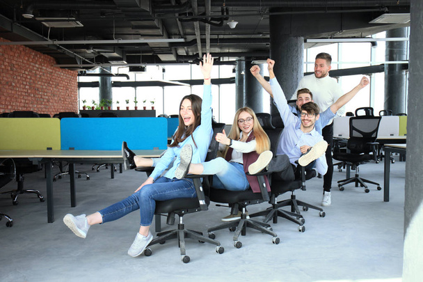 Joven gente de negocios alegre en ropa casual inteligente divirtiéndose mientras compite en sillas de oficina y sonriendo
. - Foto, Imagen