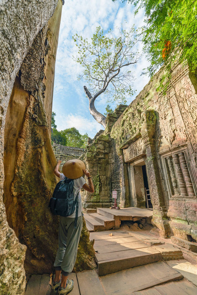 One tourist visiting Angkor ruins amid jungle, Angkor Wat temple complex, travel destination Cambodia. Woman with traditional hat, rear view. - Photo, Image