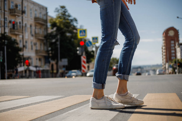 Young girl dressed in jeans and sneakers is crossing the road with a backpack in a city street on a summer sunny day - Foto, imagen