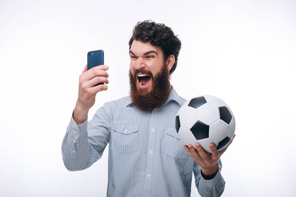 Кричащий бородатый мужчина в случайном глядя на смартфон и холдин
 - Фото, изображение