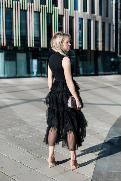 ein schönes blondes Mädchen steht mit dem Rücken in einem schwarzen Abendkleid vor der Kulisse eines modernen Gebäudes. - Foto, Bild