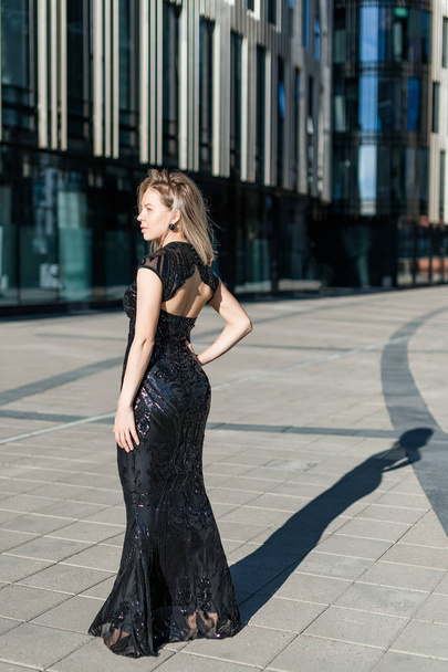 A beautiful blonde girl stands with her back in a black evening long dress against the backdrop of a modern building. - Foto, imagen
