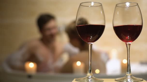 Red wine glasses standing on background of beloved couple enjoying in bathtub - Footage, Video