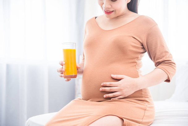 Mulher grávida sentada na cama, segurando um copo de suco de laranja e sorrisos
 - Foto, Imagem