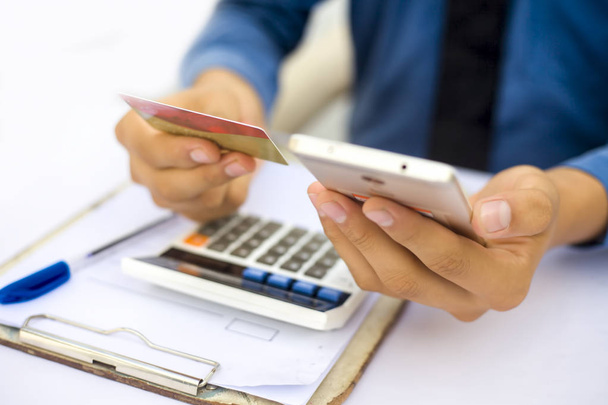 Close up of employee wearing shirt and necktie holding smartphone and credit card in hands while sitting at desk. Concept of banking and shopping  - Photo, Image