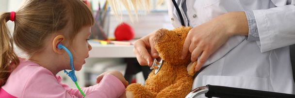 小さな女の子が腕のおもちゃの聴診器でホールドテディベアと遊ぶ - 写真・画像