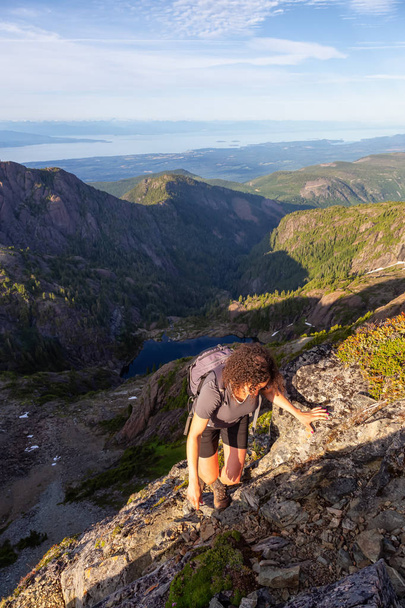 Adventurous girl climbing up a steep cliff in the Canadian Mountain Landscape during a vibrant summer evening. Taken at Mt Arrowsmith, near Nanaimo, Vancouver Island, BC, Canada. - Foto, Imagen