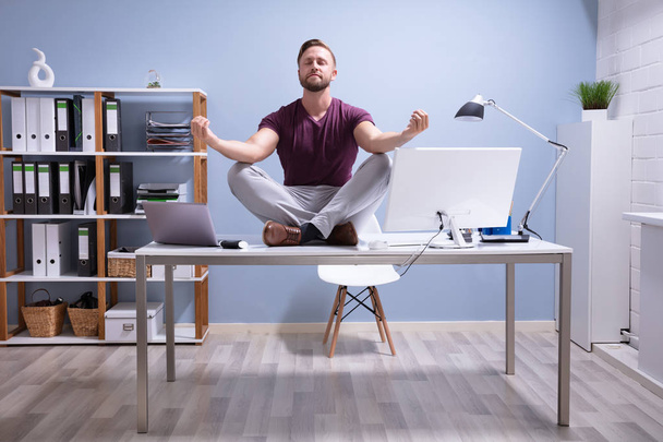 Young Businessman Sitting On Desk Meditating In Office - Foto, afbeelding