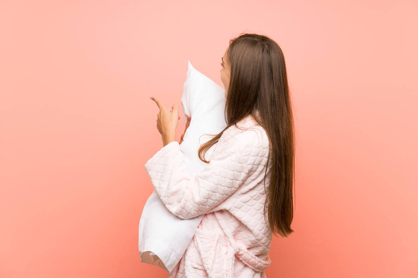 Молодая женщина в халате поверх розовой стены указывает назад указательным пальцем
 - Фото, изображение