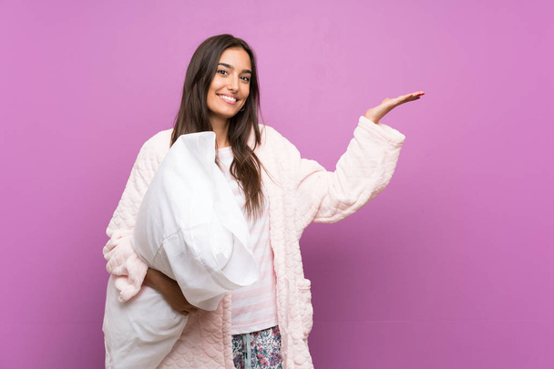junge Frau im Schlafanzug und Morgenmantel über isoliertem lila Hintergrund, die Hände zur Seite ausstreckend, um zum Kommen einzuladen - Foto, Bild