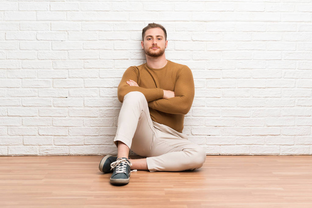 Blonde man zittend op de vloer houden armen gekruist - Foto, afbeelding