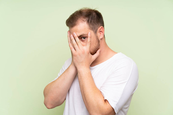 Handsome man over green background covering eyes and looking through fingers - Fotografie, Obrázek