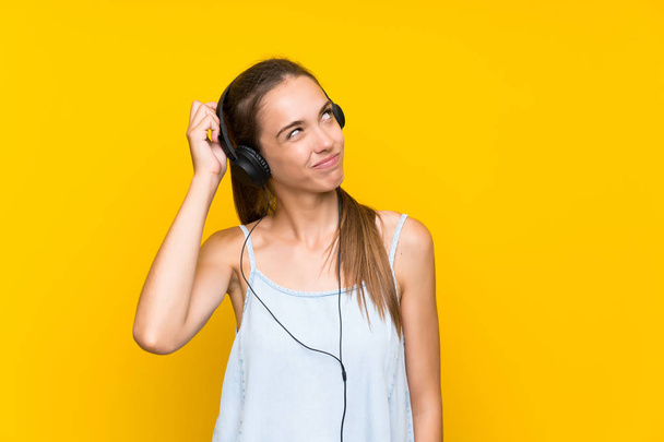 孤立した黄色い壁の上で音楽を聴いている若い女性は疑問を持ち、顔の表情を混乱させる - 写真・画像