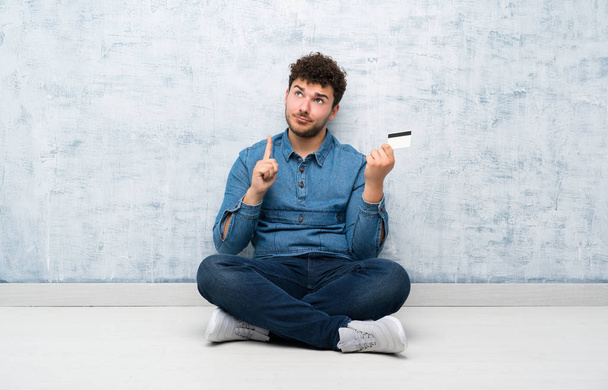 Νέος άνθρωπος κάθεται στο πάτωμα κρατώντας μια πιστωτική κάρτα - Φωτογραφία, εικόνα