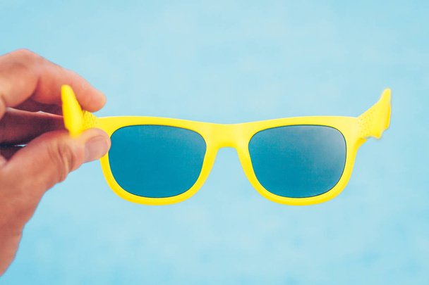 Yellow sunglasses in hand on blue sky background - 写真・画像