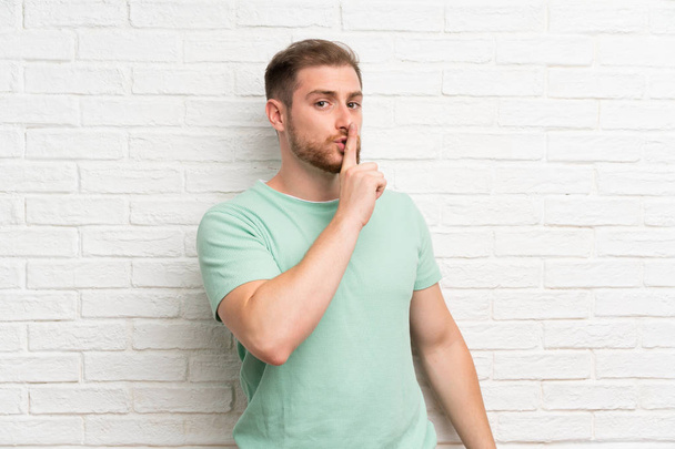 Blonde man over brick wall doing silence gesture - Foto, afbeelding