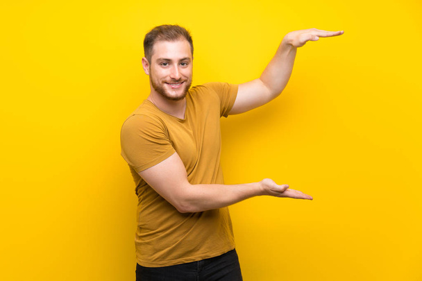 Ξανθός άνθρωπος πάνω από απομονωμένο κίτρινο τοίχο κρατώντας χώρο αντιγραφής για να εισαγάγετε μια διαφήμιση - Φωτογραφία, εικόνα