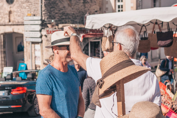 Gordes, Provence-Alpes-Cote d'Azur, France, September 25, 2018: Male tourists measure and bargain for a hat - a traditional farmers market in Provence - Fotó, kép