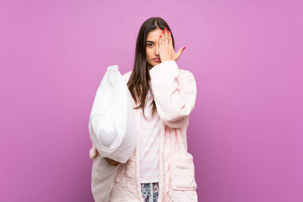 Νεαρή γυναίκα με πιτζάμες και ρόμπα πάνω από απομονωμένο μωβ φόντο που καλύπτει ένα μάτι με το χέρι - Φωτογραφία, εικόνα