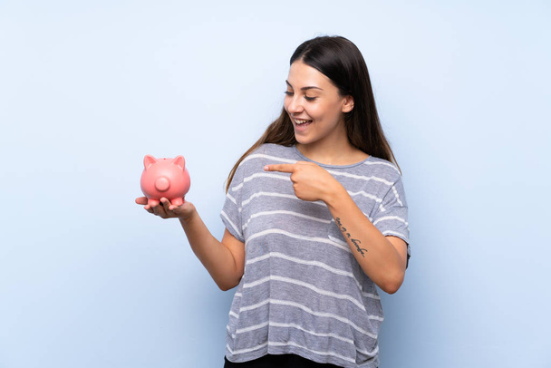 Young brunette woman over isolated blue background holding a big piggybank - Photo, Image