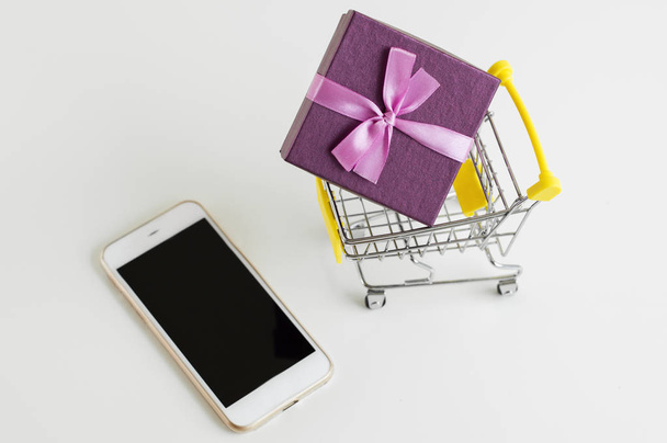 Small trolley toy with a gift box and mobile phone. Shopping with a smartphone. Online and internet shopping concept - Fotó, kép