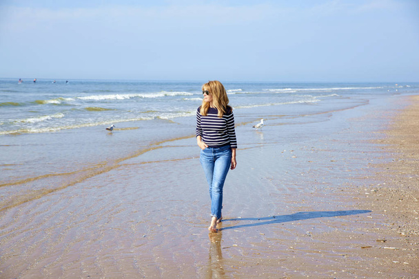 Full length shot of barefoot woman wearing casual clothes and sunglasses while walking on the seaside. - Valokuva, kuva