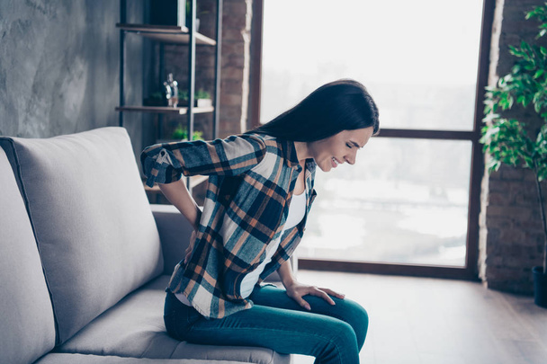 Close up side profile photo beautiful she her lady arms hands hold back spine suffering terrible loins pain wear jeans denim checkered plaid shirt sit divan comfort flat house living room indoors - Фото, изображение