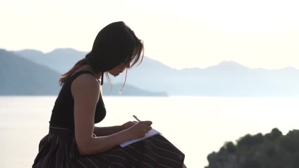 Csinos nő írja a notebook, ülve a tenger közelében lassítva napkelte - Felvétel, videó