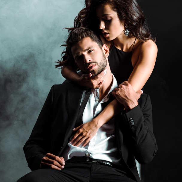 passionate woman standing and hugging handsome bearded man on black with smoke  - Photo, Image