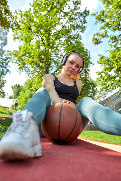 Menina bonita, atlética com uma bola de basquete escuta música com fones de ouvido. Motivação desportiva, estilo de vida saudável
. - Foto, Imagem