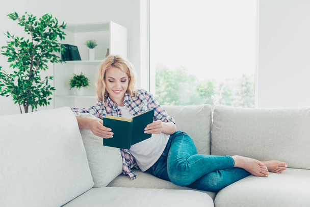 Portrait of her she nice-looking winsome attractive lovely charming cute cheerful cheery wavy-haired girl in checked shirt sitting on divan reading novel at white light style interior house room - Zdjęcie, obraz