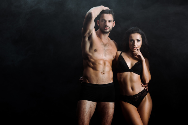 handsome shirtless man standing and touching hair near attractive girl in lingerie on black with smoke  - Photo, Image